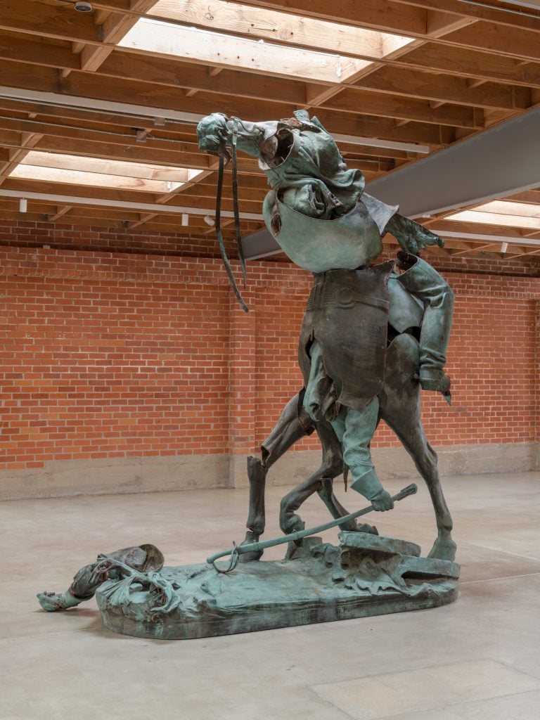 A photo shows a large art gallery with a brick wall. At its center stands an 11-foot-high bronze artwork created by rearranging the parts of an equestrian monument