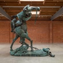 A photo shows a large art gallery with a brick wall. At its center stands an 11-foot-high bronze artwork created by rearranging the parts of an equestrian monument