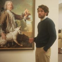 A white man with dark hair stands in an art museum gallery, with paintings on the wall, wearing khakis and a green sweater