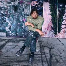 The artist Kristian Touborg sits on a low stool in a paint-splattered studio, surrounded by large abstract canvases featuring swirling, water-like patterns in deep purples, blues, and greens. He wears paint-streaked clothes and black sneakers, gazing directly at the camera with an intense expression. The floor beneath him is also covered in marks and drips of paint, adding to the creative, immersive atmosphere.