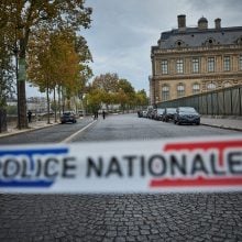 Police tape reading “Police Nationale” blocks cobblestone street outside Louvre, with investigators visible in distance.