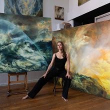 Maria Kreyn in a black outfit sits barefoot on a stool in an art studio, surrounded by large, dramatic abstract landscape paintings. The artworks feature swirling, cloud-like forms in deep blue-green tones on one side and warm golden-yellow tones on the other. The setting is a spacious studio with wooden floors and canvases leaning against the walls.