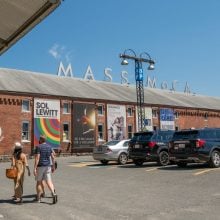 Three visitors walk toward MASS MoCA’s brick exterior, featuring large banners advertising contemporary art exhibitions.