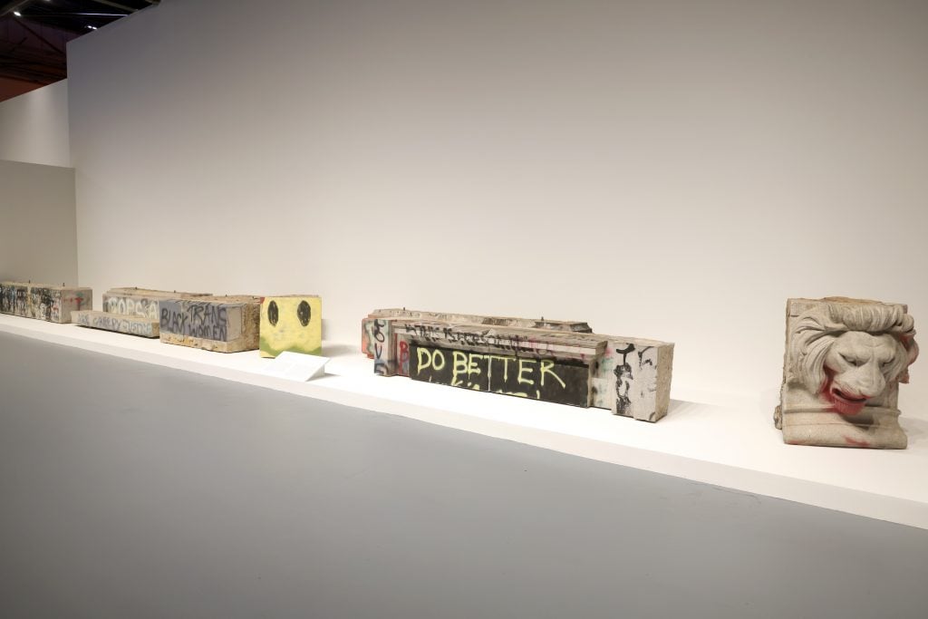 Contemporary gallery installation of graffitied architectural fragments and protest-marked stone blocks displayed in a minimalist white exhibition space.
