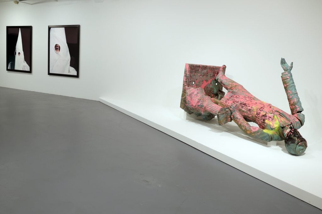 A photo shows a toppled bronze monument of a man, lying on its side, covered in pink paint, and on the wall nearby photos of men in Ku Klux Klan hoods