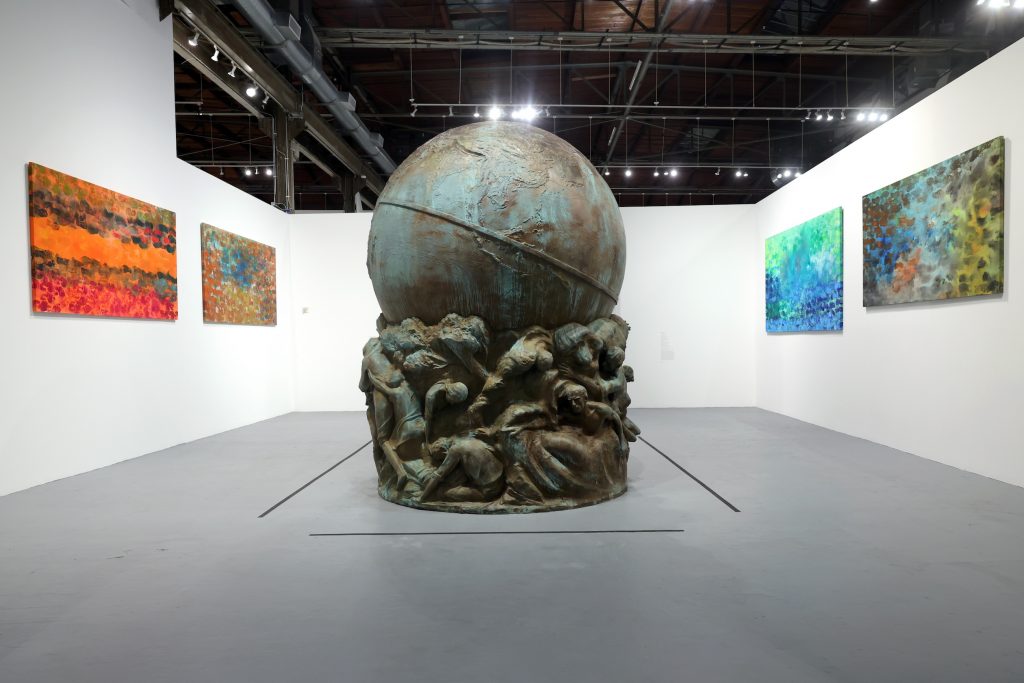 A photo shows an art gallery with abstract paintings on the wall surrounding a large bronze monument in the shape of a globe.