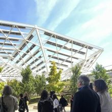 At Museums of Tomorrow, a group of participants listen to speaker under striking solar canopy amid trees and blue sky.