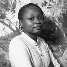 Black-and-white portrait of Ng'onga Silupya of Imvelo Studios standing with arms crossed in front of an abstract, textured painting. She wears a light-colored button-up shirt and looks confidently at the camera. The background features expressive brushstrokes and splatters, adding a dynamic, artistic atmosphere to the composition.