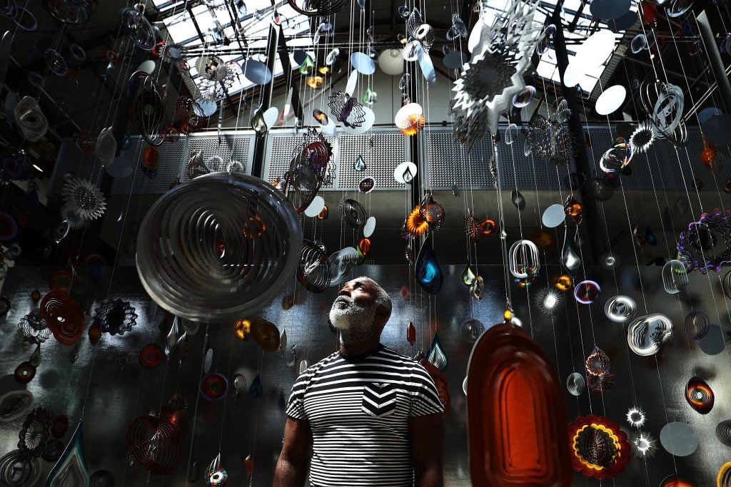 Nick Cave standing beneath a dazzling installation of suspended, reflective, and colorful kinetic sculptures that hang from the ceiling in a skylit gallery, gazing upward amid swirling light and shadow.
