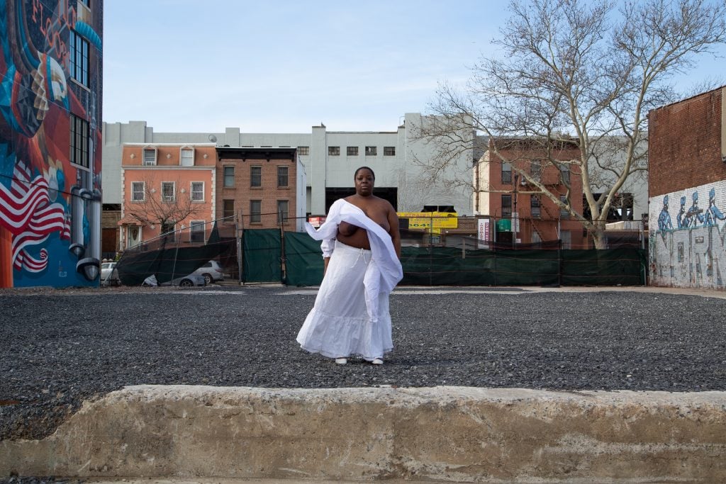 An artwork in the form of a photograph shows a Black woman, the artist Nona Faustine, standing in a vacant lot in Brooklyn, with buildings all around. She stands alone, partially clothed in flowing white garments, parts of her upper body exposed