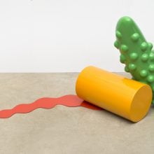 A sculpture shows a green form resembling a pickle leaning on a yellow cylinder resting on the floor, from under which comes a red puddle-like form