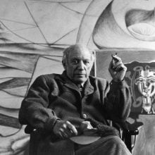 A black-and-white photograph of Pablo Picasso seated in his studio, holding a cigarette, with large abstract paintings visible behind him.