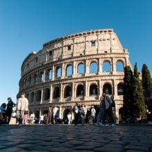 Tourists gather around the ancient Roman Colosseum under a clear blue sky, its arches bathed in sunlight.