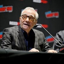 Film industry panel discussion at a convention, with a speaker addressing the audience beside another participant wearing sunglasses.