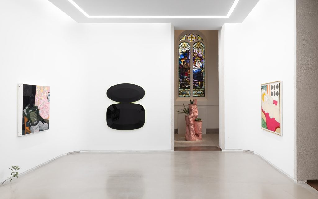 A gallery space with white walls and polished floors featuring several artworks, including a seated figure painting, two glossy black wall sculptures, and a colorful abstract piece. A pink sculptural installation and stained-glass window are visible through an arched doorway.