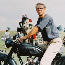 A photo of a light-skinned man in his 30s or 40s with short blond hair wearing a dark blue T-shirt and beige pants, sitting on a black motorcycle outdoors with people and film equipment in the background.