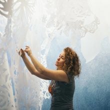 A photograph shows a woman, artist Swoon, against the backdrop of one of her artworks