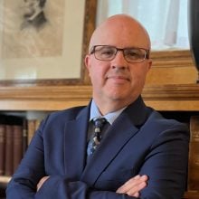 Professional man in suit and glasses posing with arms crossed in office library setting.