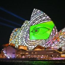 The Sydney Opera House illuminated at night with vivid projections, featuring a large green eye surrounded by colorful mosaic patterns during a light festival.