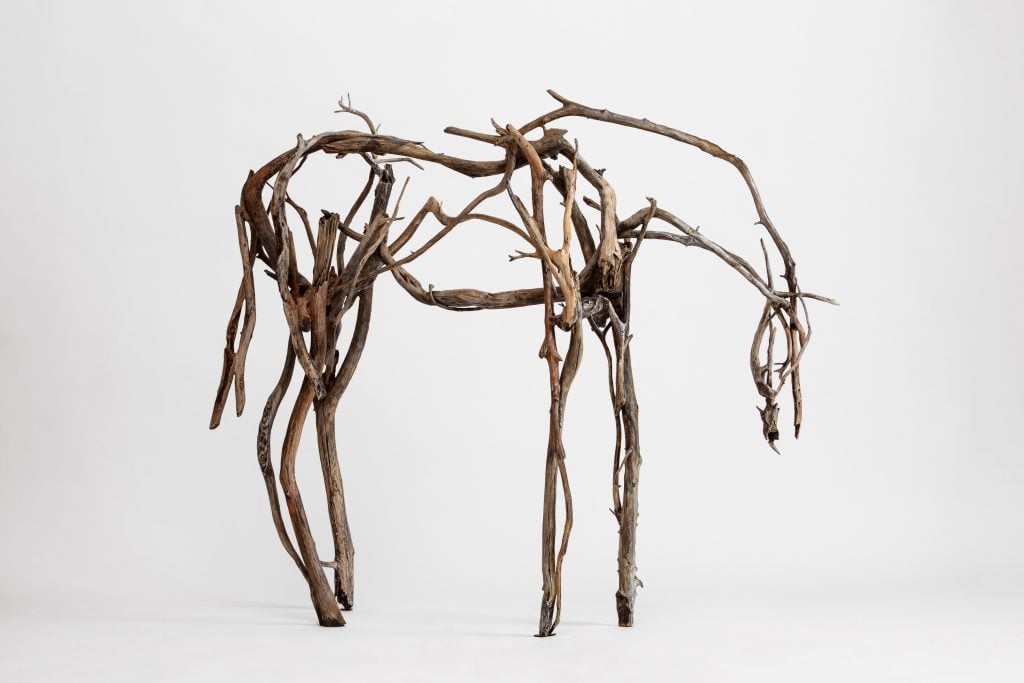 A life-sized horse sculpture constructed from intertwined pieces of driftwood, standing against a plain white background. The organic branches form a delicate yet dynamic outline of the horse’s body, legs, and head.