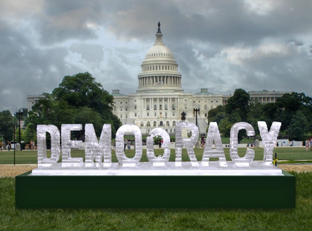 A photo of a large ice sculpture spelling “DEMOCRACY” on the National Mall, with the U.S. Capitol building rising behind it under a cloudy sky and distant visitors scattered across the lawn.