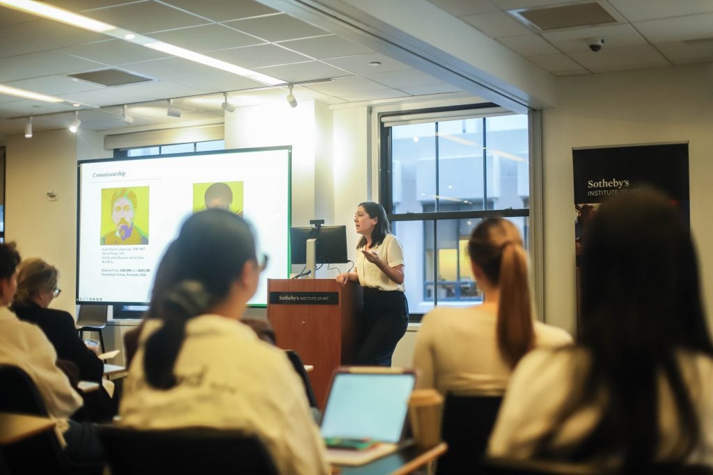 Kait McElwee of Arcadia Art Consultancy gives a lecture at the Sotheby’s Institute of Art, standing beside a podium and gesturing toward a projected slide that shows two Andy Warhol portraits. Several attendees are seated in the classroom, listening and taking notes on laptops.