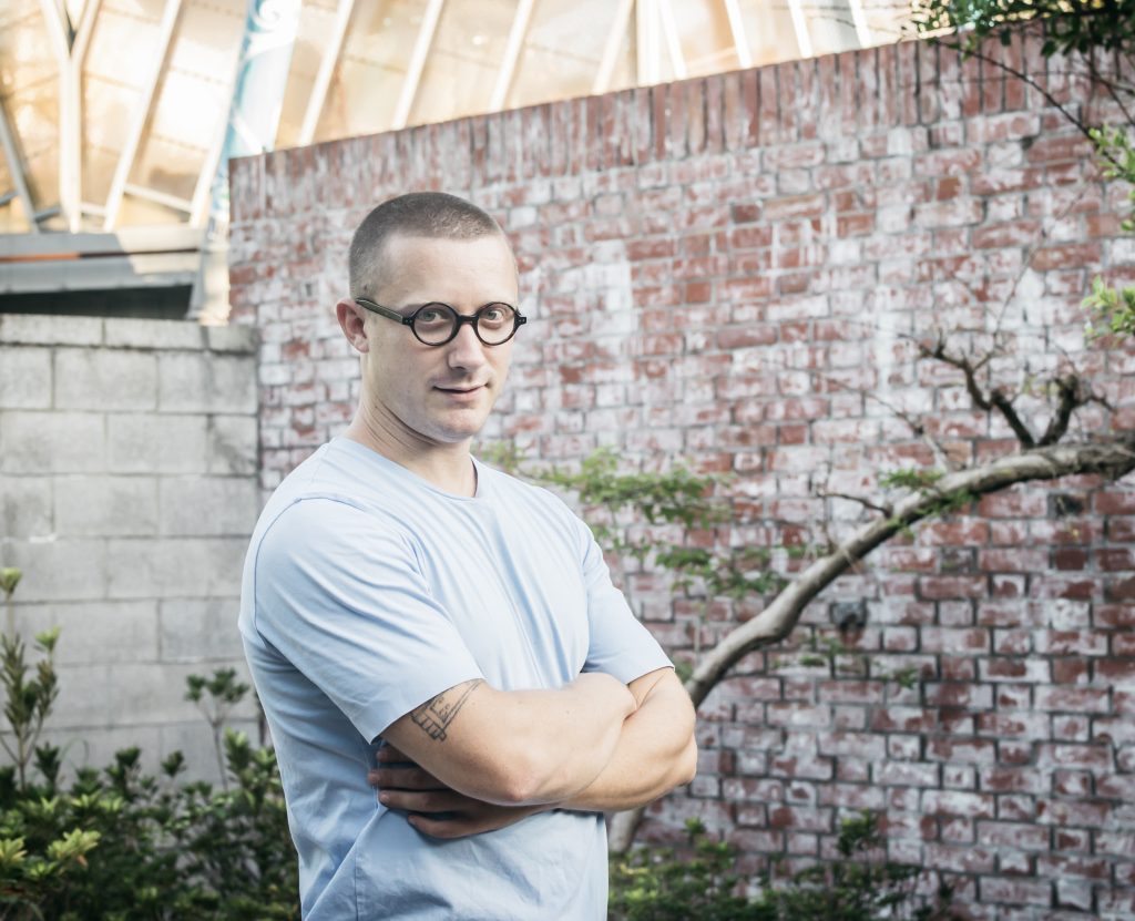 photo of a bespectacled caucasian man skin head in baby blue T-shirt, standing in front of a faded red brick wall.